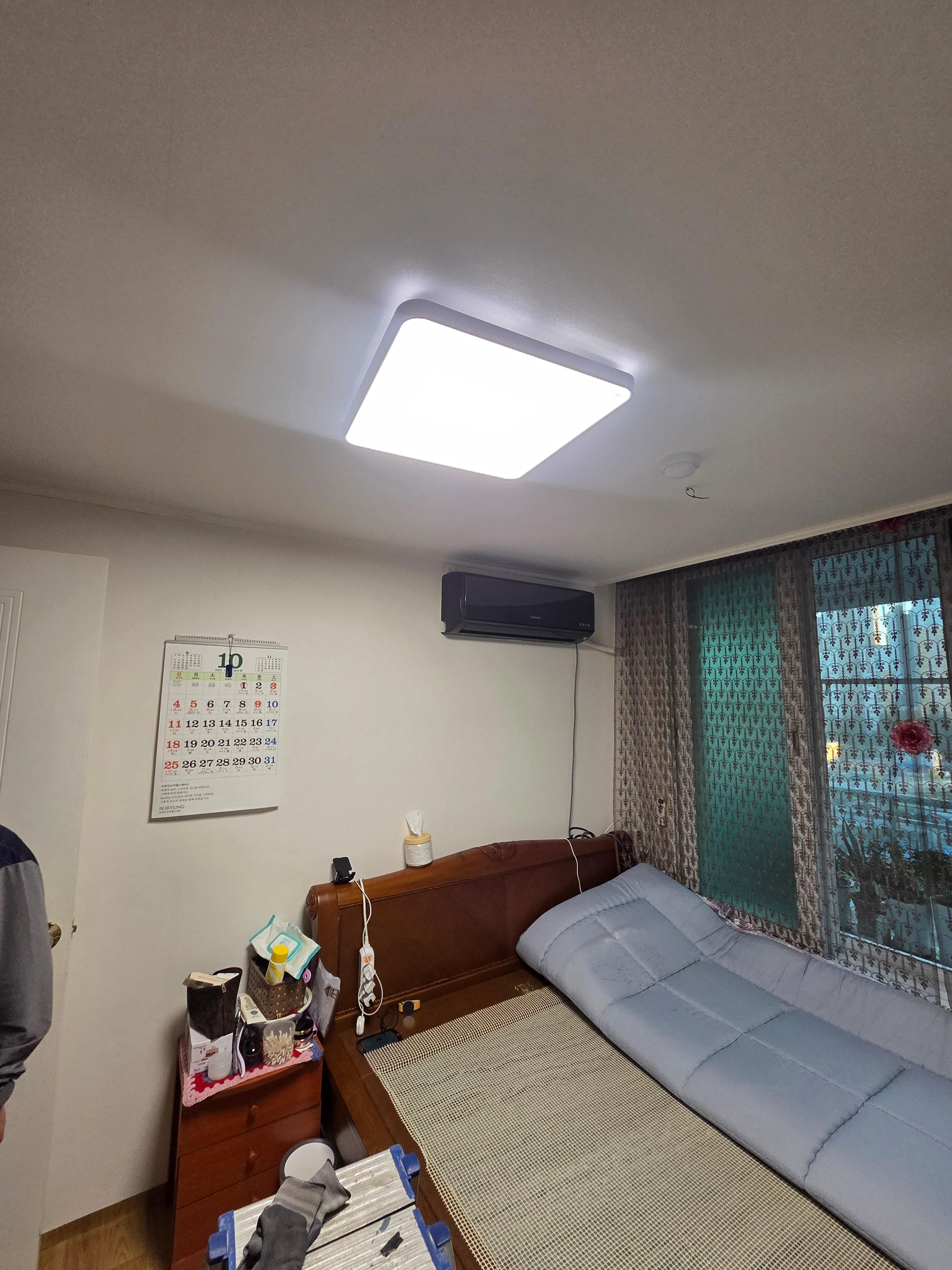 침대와 벽걸이 에어컨이 있는 방의 천장에 설치되어 켜진 사각 조명